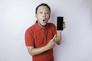 Surprised Asian man wearing red t-shirt pointing at his smartphone, isolated by white background photo