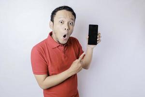 hombre asiático sorprendido con una camiseta roja apuntando a su teléfono inteligente, aislado de fondo blanco foto