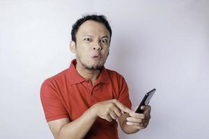An angry young Asian man looks disgruntled wearing red shirt irritated face expressions holding his phone photo