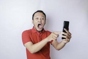 Surprised Asian man wearing red t-shirt pointing at his smartphone, isolated by white background photo