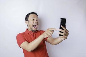 Surprised Asian man wearing red t-shirt pointing at his smartphone, isolated by white background photo