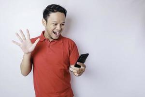hombre asiático sorprendido con una camiseta roja apuntando a su teléfono inteligente, aislado de fondo blanco foto