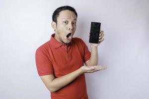 Surprised Asian man wearing red t-shirt pointing at his smartphone, isolated by white background photo