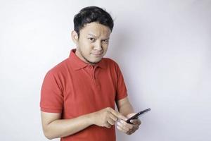 Portrait of a thoughtful young Asian man wearing red t-shirt looking aside while holding smartphone photo