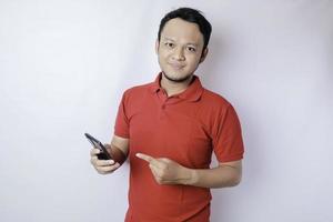 A portrait of a happy Asian man is smiling and holding his smartphone wearing a red t-shirt isolated by a white background photo