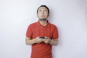 A dissatisfied young Asian man looks disgruntled wearing red t-shirt irritated face expressions holding his phone photo