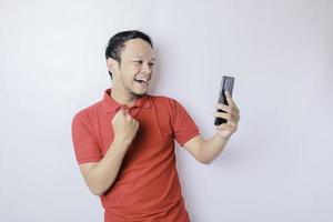 A young Asian man with a happy successful expression wearing red shirt and holding smartphone isolated by white background photo