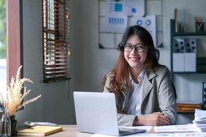 Pretty Asian woman sitting in the office, calculator, working on data, charts and documents on the table in business workspace. Looking camera. photo