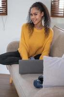 una mujer afroamericana usa una computadora portátil, un teléfono inteligente, una tableta y un auricular de música en su casa para relajarse. foto