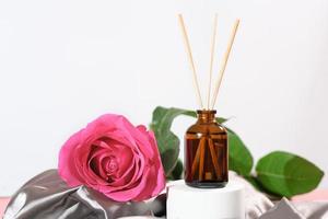 home aroma diffuser with reed sticks, rose flower scent. cozy home, romantic ambience, silk satin bedding sheets. pink rose flower next to glass bottle with fragranced oil. photo