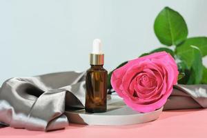 Product for skincare based on rose oil, essential oil solution ina dropper bottle, brown pipette bottle and fresh pink rose flower next to silk textile cloth. photo