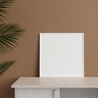 Minimalist front view square white photo or poster frame mockup leaning against wall on table with plant. 3d rendering.