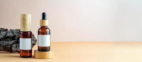 A mock-up of a brown cosmetics bottles with dispenser and white label and dropper bottle with pipette on wooden podium for cosmetics photography on beige background. Banner photo