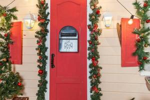 Christmas porch decoration idea. House entrance with red door decorated for holidays. Red and green wreath garland of fir tree branches and lights on railing. Christmas eve at home photo