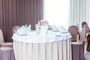 mesa elegante para cenar con composición floral en el restaurante, fondo interior de lujo. boda elegante decoración de banquetes y artículos para comida organizados por servicio de catering en mesa blanca. foto