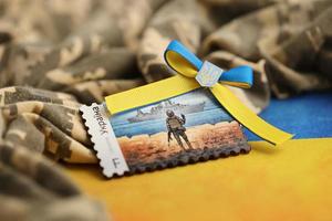 TERNOPIL, UKRAINE - SEPTEMBER 2, 2022 Famous Ukrainian postmark with russian warship and ukrainian soldier as wooden souvenir on army camouflage uniform and national flag photo