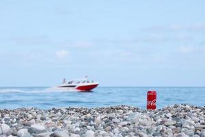 ANTALYA, TURKEY - MAY 18, 2021 Original Coca Cola red tin can lies on small round pebble stones close to sea shore. Coca-cola can and speed boat on beach photo