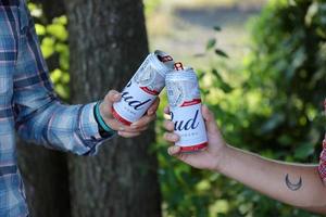 SUMY, UKRAINE - AUGUST 01, 2021 Young man raise Budweiser Bud beer can with male friend on blurred river with kayak and trees. Budweiser is one of the most popular beer brands in the USA photo