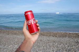 ANTALYA, TURKEY - MAY 18, 2021 Original Coca Cola red tin can lies on small round pebble stones close to sea shore. Coca-cola can in male hand on beach photo