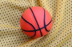 Small orange rubber basketball lies on a yellow sport jersey clothing fabric texture and background with many folds photo