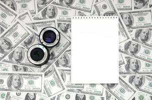 Two photographic lenses and white notebook lie on the background of a lot of dollar bills. Space for text photo