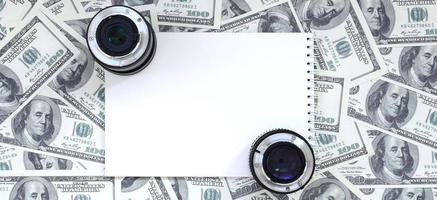 dos lentes fotográficos y un cuaderno blanco se encuentran en el fondo de muchos billetes de dólar. espacio para texto foto