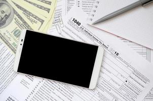 Composition of items lying on the 1040 tax form. Dollar bills, pen, smartphone, paper clip and notepad. Clean black screen smartphone for text placement photo