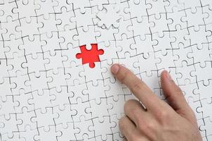 The texture of a white jigsaw puzzle in the assembled state with one missing element, forming a red space, pointed to by the finger of the male hand photo