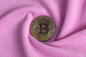 The golden bitcoin lies on a blanket made of soft and fluffy light pink fleece fabric with a large number of relief folds. The shape of the folds resembles a fan from a video card cooler photo