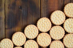 A round sandwich cookie with coconut filling lies in large quantities on a brown wooden surface. Photo of edible treats on a wooden background with copy space