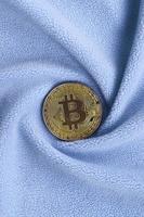 The golden bitcoin lies on a blanket made of soft and fluffy light blue fleece fabric with a large number of relief folds. The shape of the folds resembles a fan from a video card cooler photo