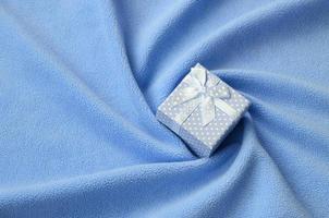 A small gift box in blue with a small bow lies on a blanket of soft and furry light blue fleece fabric with a lot of relief folds. Packing for a gift to your lovely girlfriend photo