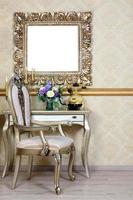 A fragment of a retro interior with a chair and table, on which is a telephone and a vase of flowers. Above the table hangs an empty picture frame for your photo