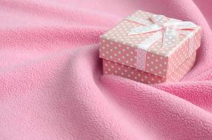 A small gift box in pink with a small bow lies on a blanket of soft and furry light pink fleece fabric with a lot of relief folds. Packing for a gift to your lovely girlfriend photo