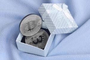 The silver bitcoin lies in a small blue gift box with a small bow on a blanket made of soft and fluffy light blue fleece fabric with a large number of relief folds photo