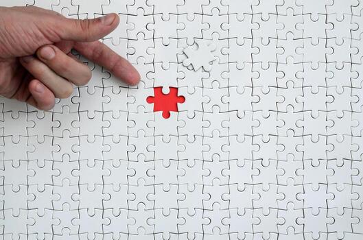 The texture of a white jigsaw puzzle in the assembled state with one missing element, forming a red space, pointed to by the finger of the male hand photo