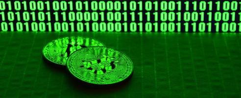 A pair of bitcoins lies on a cardboard surface on the background of a monitor depicting a binary code of bright green zeros and one units on a black background. Low key lighting photo