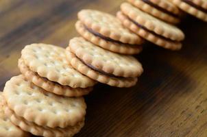 A round sandwich cookie with coconut filling lies in large quantities on a brown wooden surface. Photo of edible treats on a wooden background with copy space