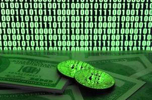 Two bitcoins lies on a pile of dollar bills on the background of a monitor depicting a binary code of bright green zeros and one units on a black background. Low key lighting photo