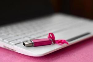 la tarjeta de memoria flash usb de color rosa brillante con un lazo rosa se encuentra sobre una manta de tela suave y peluda de color rosa claro junto a una computadora portátil blanca. diseño clásico de regalo femenino para una tarjeta de memoria foto