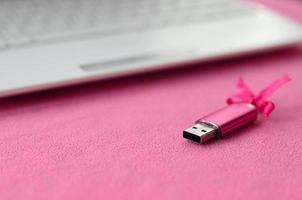 Brilliant pink usb flash memory card with a pink bow lies on a blanket of soft and furry light pink fleece fabric beside to a white laptop. Classic female gift design for a memory card photo