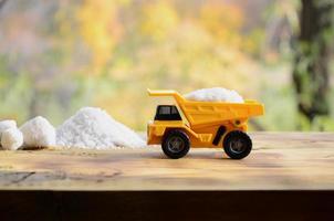 A small yellow toy truck is loaded with a stone of white salt next to a pile of salt. A car on a wooden surface against a background of autumn forest. Extraction and transportation of salt photo