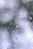 A photo of rain drops on the window glass with a blurred view of the blossoming green trees. Abstract image showing cloudy and rainy weather conditions