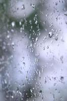 A photo of rain drops on the window glass with a blurred view of the blossoming green trees. Abstract image showing cloudy and rainy weather conditions
