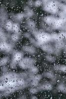 A photo of rain drops on the window glass with a blurred view of the blossoming green trees. Abstract image showing cloudy and rainy weather conditions