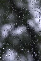 A photo of rain drops on the window glass with a blurred view of the blossoming green trees. Abstract image showing cloudy and rainy weather conditions