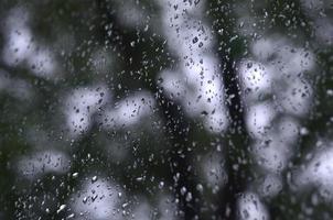 A photo of rain drops on the window glass with a blurred view of the blossoming green trees. Abstract image showing cloudy and rainy weather conditions