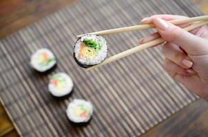 A hand with chopsticks holds a sushi roll on a bamboo straw serwing mat background. Traditional Asian food photo