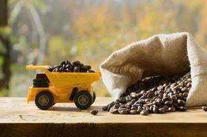 A small yellow toy truck is loaded with brown coffee beans around a full bag of grains. A car on a wooden surface against a background of autumn forest. Extraction and transportation of coffee photo