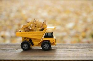 A small toy yellow truck is loaded with yellow fallen leaves. The car stands on a wooden surface against a background of a blurry autumn park. Cleaning and removal of fallen leaves. Seasonal works photo